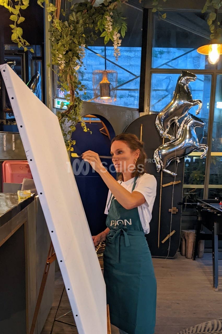 Une femme peint sur une grande toile blanche dans un espace intérieur décoré de plantes et de ballons argentés.