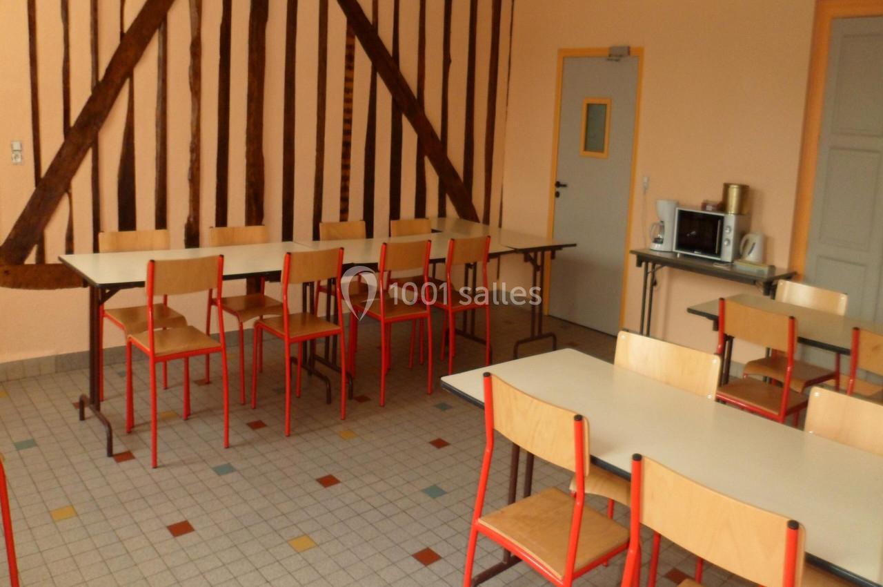 Salle avec des tables et chaises en bois, sol carrelé, murs beige avec poutres apparentes et équipements de cuisine.
