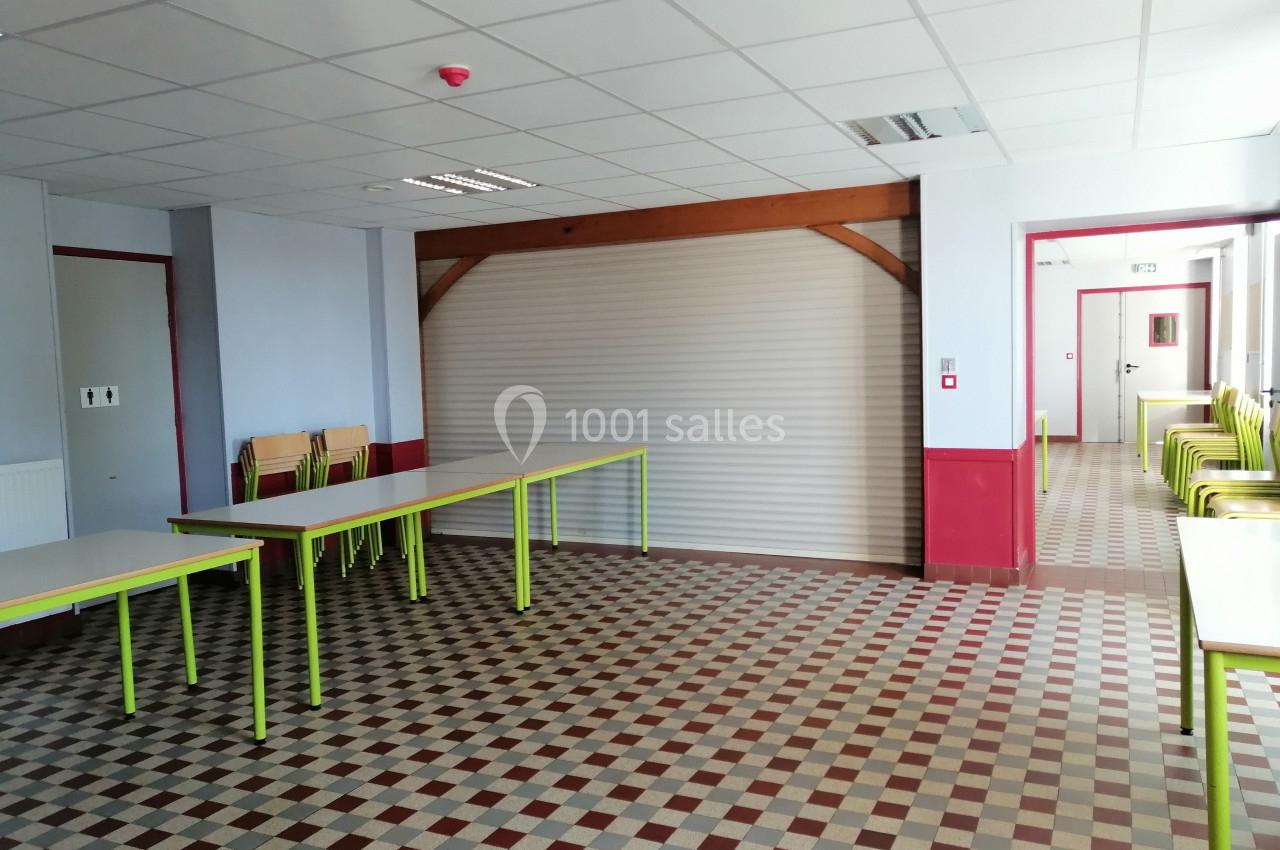 Salle vide avec tables et chaises empilées, sol à carreaux, murs clairs et rideau métallique fermé au fond.