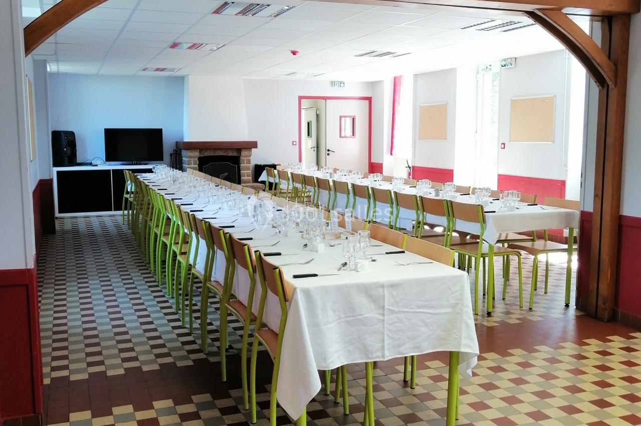 Salle lumineuse avec tables alignées, nappes blanches, chaises vertes et sol à carreaux, prête pour un événement.