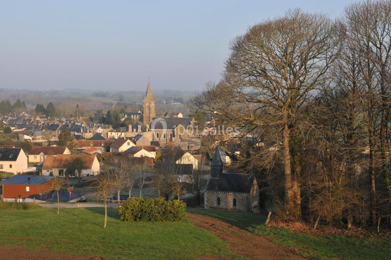 Village rural avec église au centre, maisons environnantes, arbres dénudés et paysage vallonné en arrière-plan.
