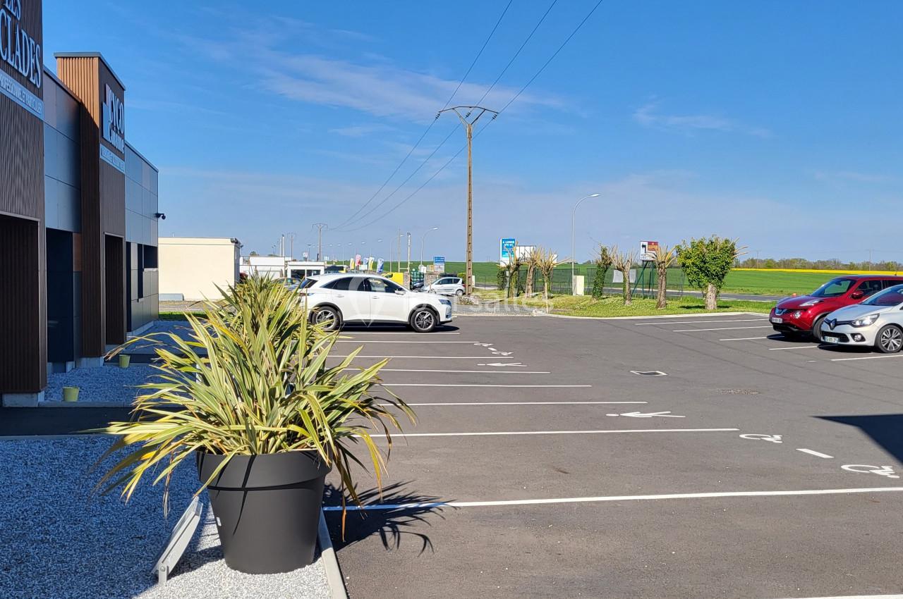 Parking extérieur avec marquages au sol, quelques voitures stationnées, bâtiments et champs en arrière-plan.