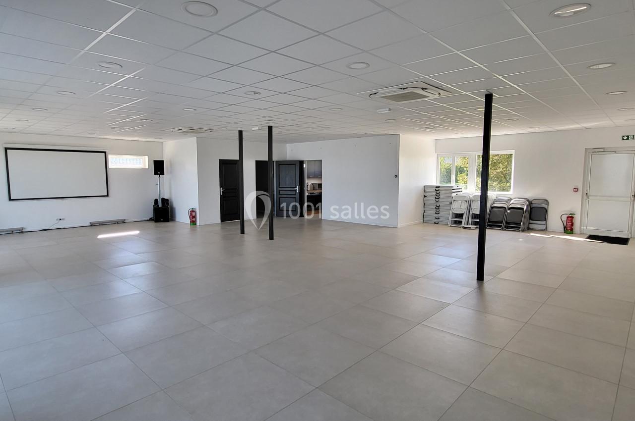 Salle vide lumineuse avec carrelage clair, murs blancs, écran de projection, chaises empilées et baies vitrées.