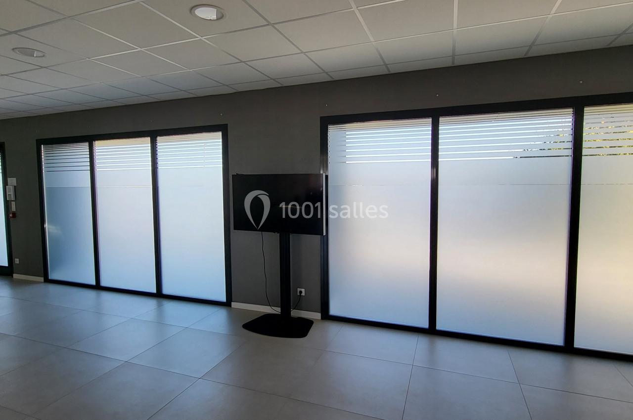 Salle lumineuse avec grandes baies vitrées partiellement opaques, sol en carrelage clair et écran sur pied.