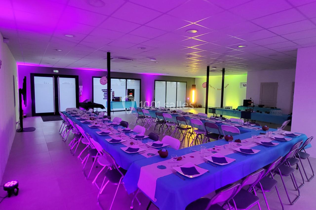 Salle de réception éclairée par des lumières colorées, tables dressées avec nappes bleues et chaises alignées.