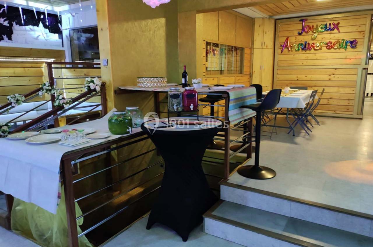 Salle décorée pour un anniversaire avec tables dressées, guirlande colorée et buffet disposé près d'un escalier.