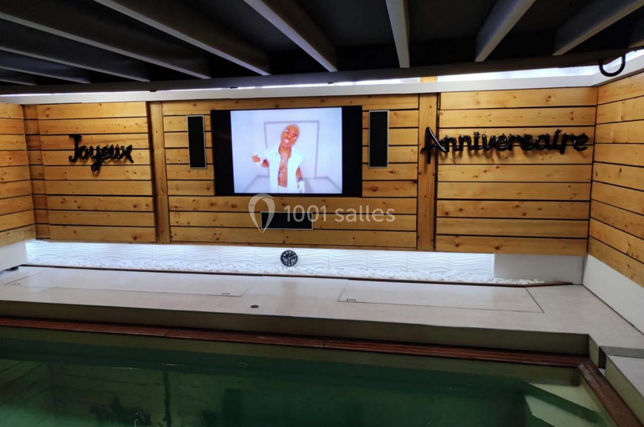 Mur en bois avec un écran affichant une personne souriante, entouré des mots ’Joyeux Anniversaire’, au-dessus d'une piscine.