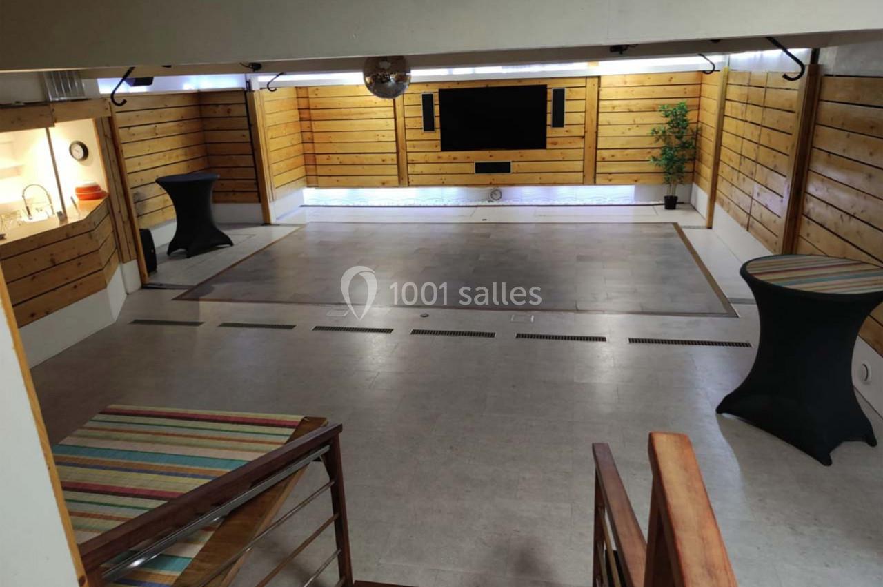 Salle polyvalente avec murs en bois, sol en carrelage, écran mural, tables hautes et coin cuisine visible à gauche.