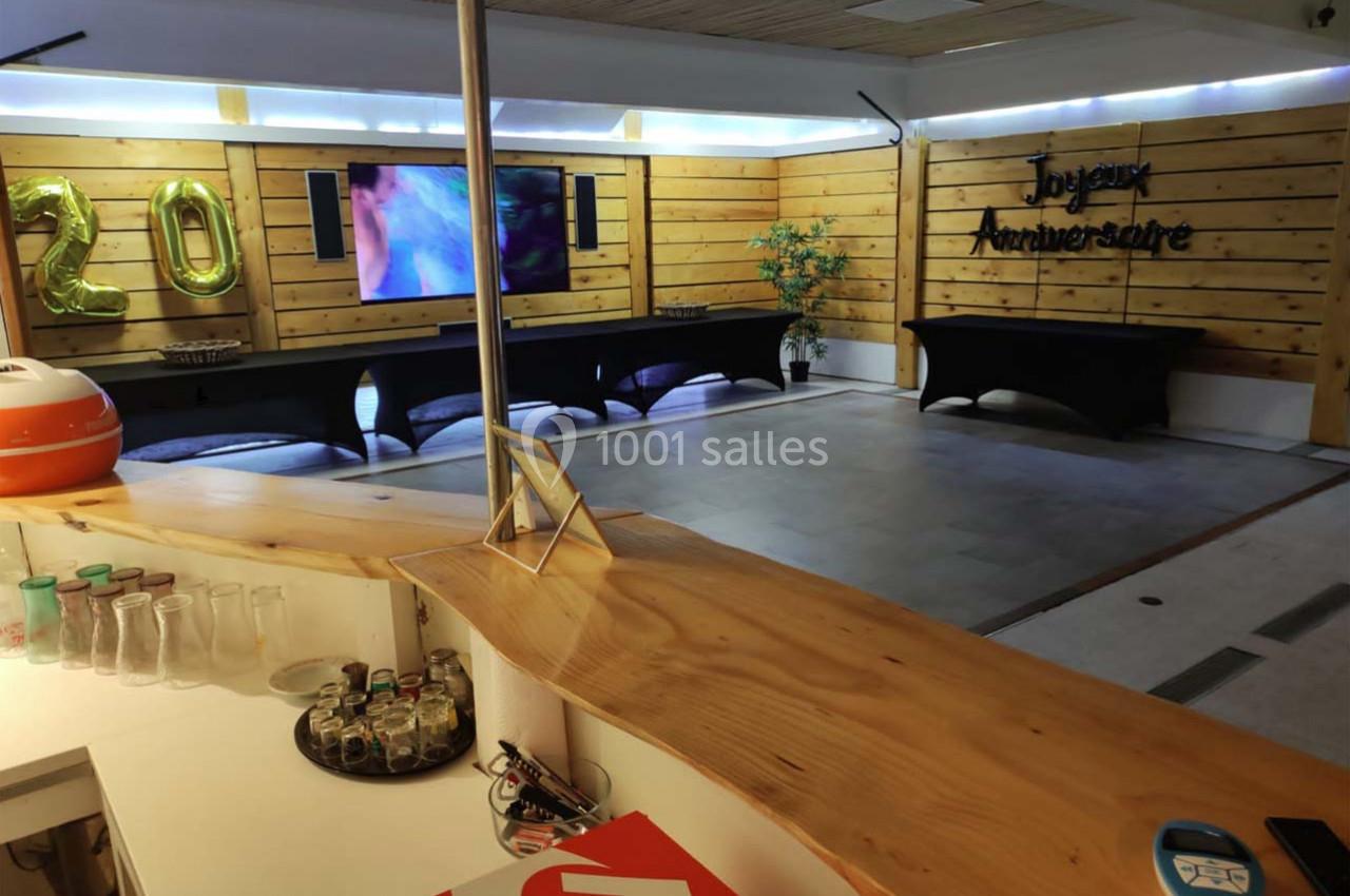 Salle décorée pour un anniversaire avec ballons dorés ’20’, écran allumé, tables noires et comptoir en bois au premier plan.