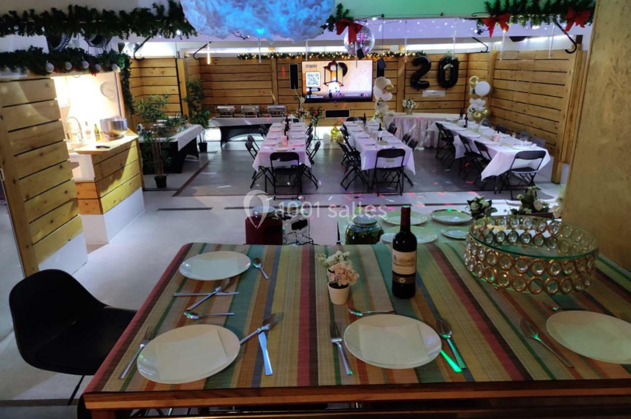 Salle décorée pour un événement avec tables dressées, guirlandes, et espace repas en arrière-plan.