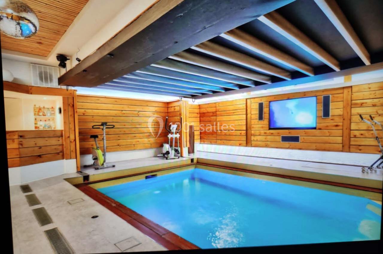 Piscine intérieure entourée de murs en bois, avec équipements de fitness et écran mural dans un espace lumineux.