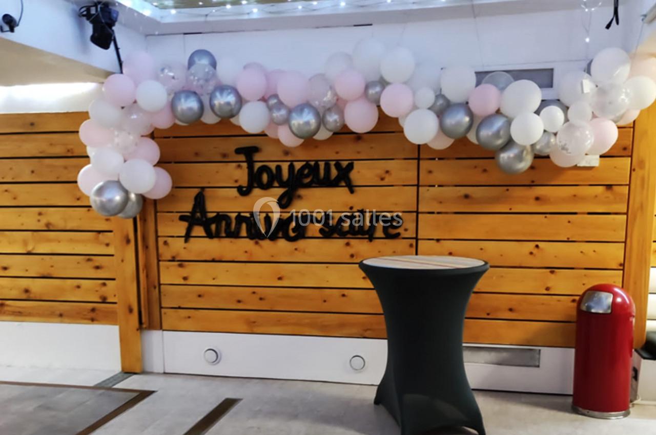 Décoration d'anniversaire avec des ballons blancs, roses et argentés formant une arche devant un mur en bois.