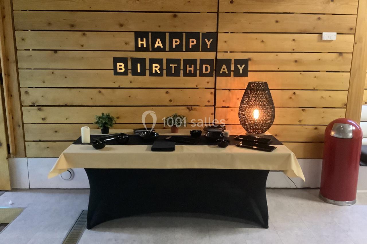 Table décorée pour un anniversaire avec vaisselle noire, nappe beige et noire, lampe allumée et panneau ’Happy Birthday’.