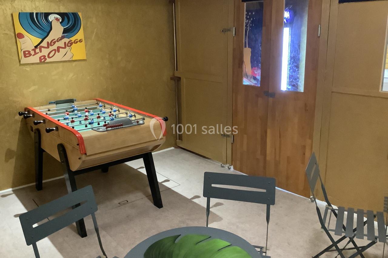 Salle avec un baby-foot en bois, une table ronde avec motif de feuille, des chaises pliantes et une affiche murale colorée.