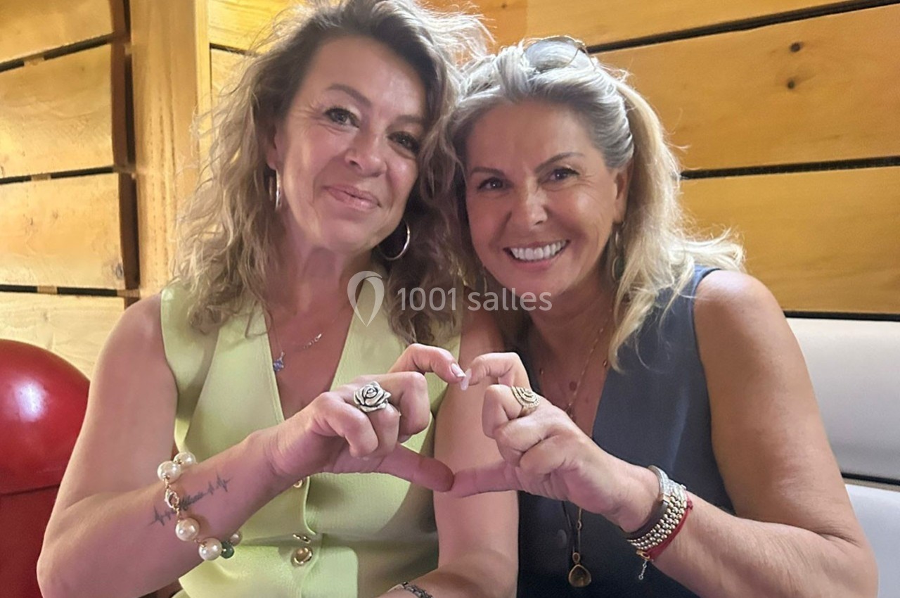 Deux femmes souriantes assises côte à côte forment un cœur avec leurs mains dans un cadre en bois.