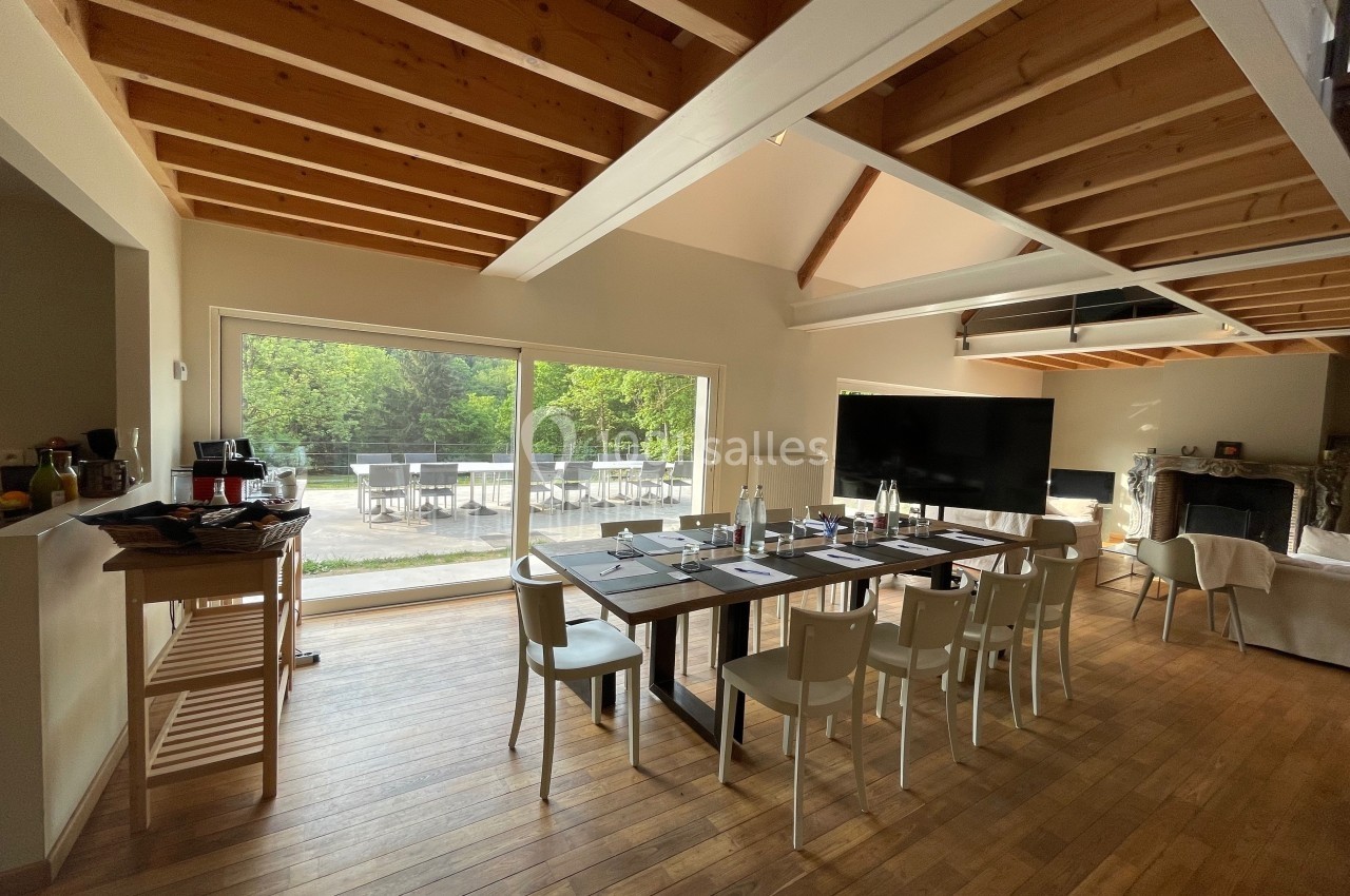 Salle lumineuse avec une grande table dressée, baies vitrées donnant sur un jardin et plafond avec poutres apparentes.