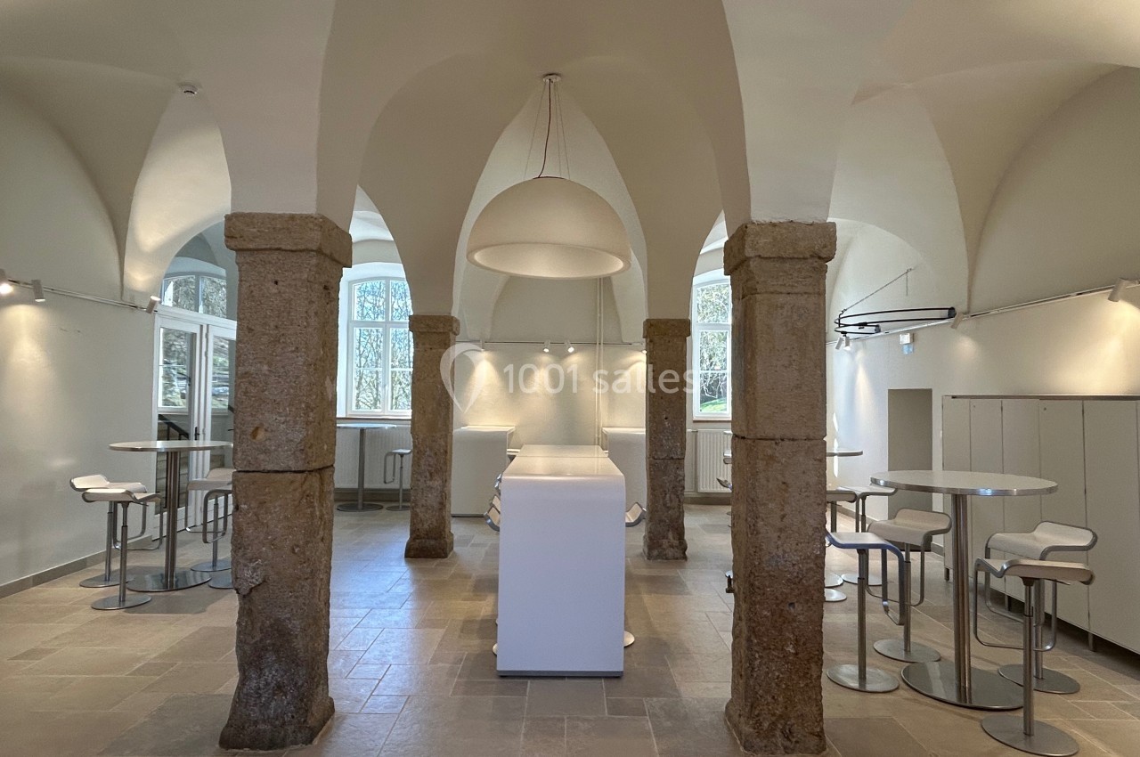 Salle voûtée lumineuse avec piliers en pierre, mobilier moderne blanc et grandes fenêtres donnant sur l'extérieur.