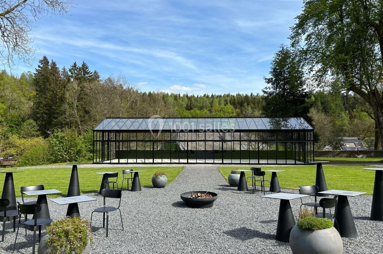 Espace extérieur avec tables, chaises et braseros, entouré de pelouse et d'arbres, avec une serre en arrière-plan.