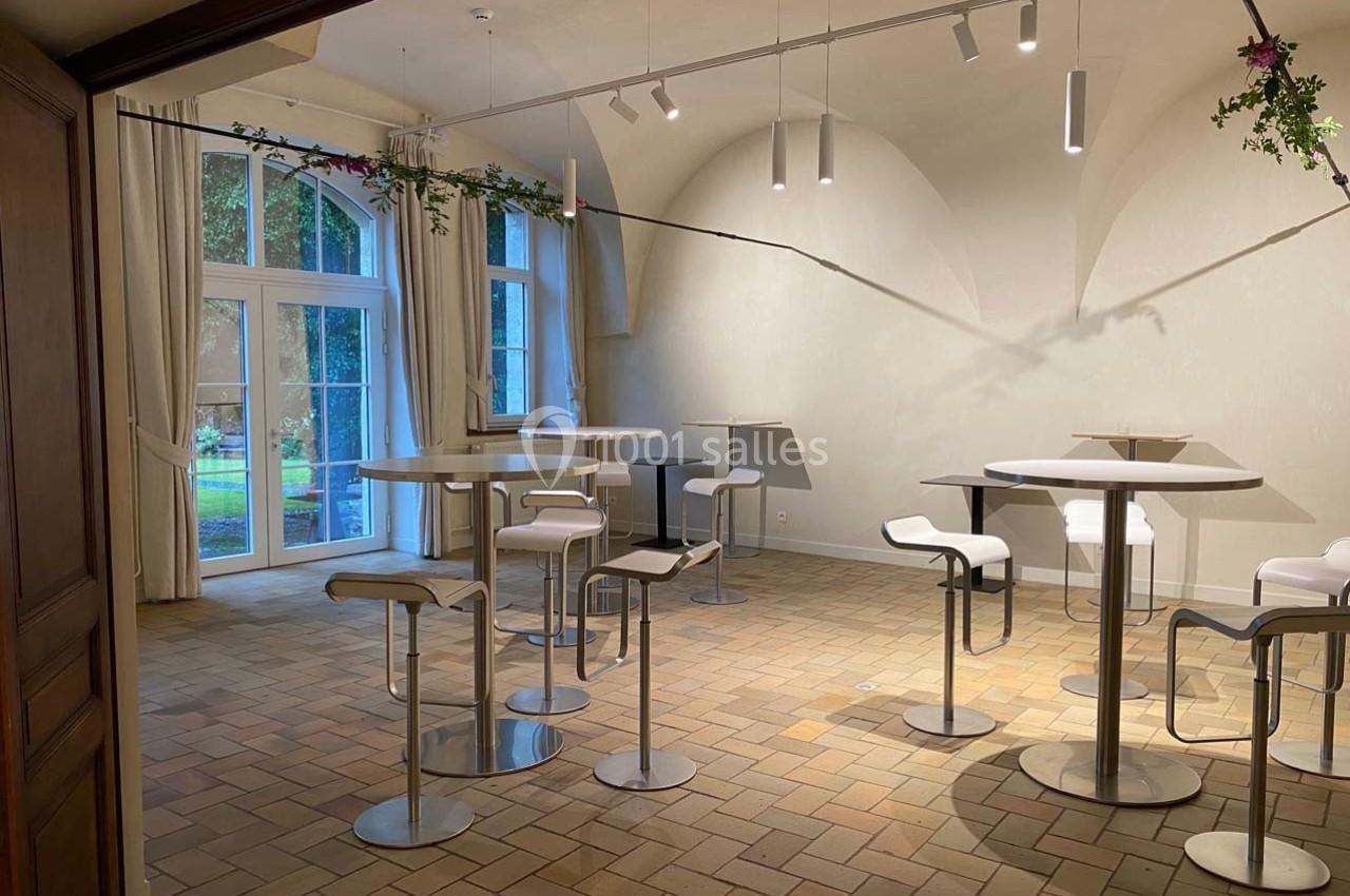 Salle lumineuse avec tables hautes, tabourets blancs et grandes fenêtres donnant sur un jardin verdoyant.