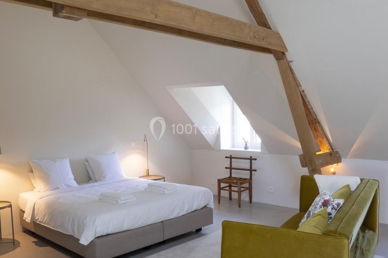 Chambre lumineuse sous combles avec lit double, poutres apparentes, canapé jaune et chaise en bois près d'une fenêtre.