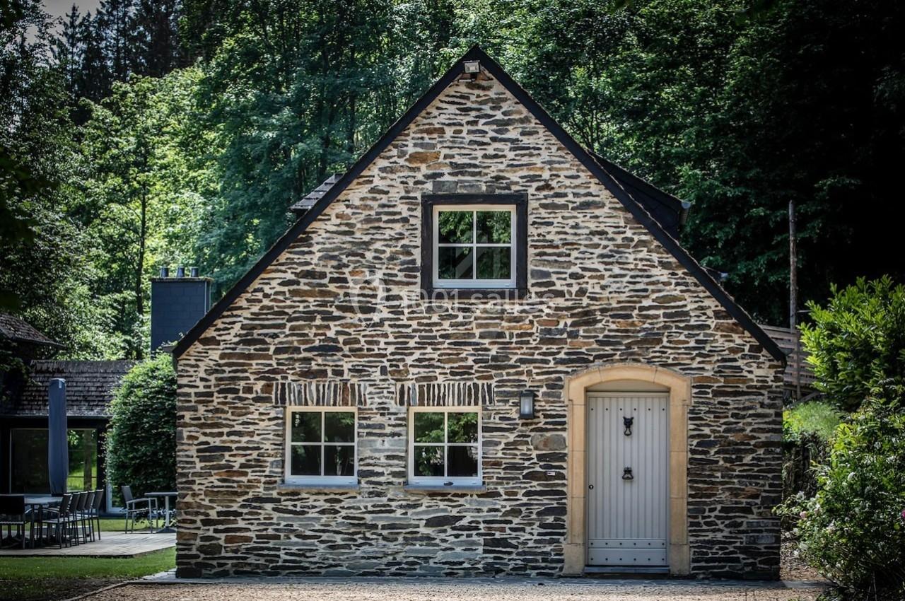 Façade d'une maison en pierre avec porte beige, fenêtres blanches et environnement boisé en arrière-plan.