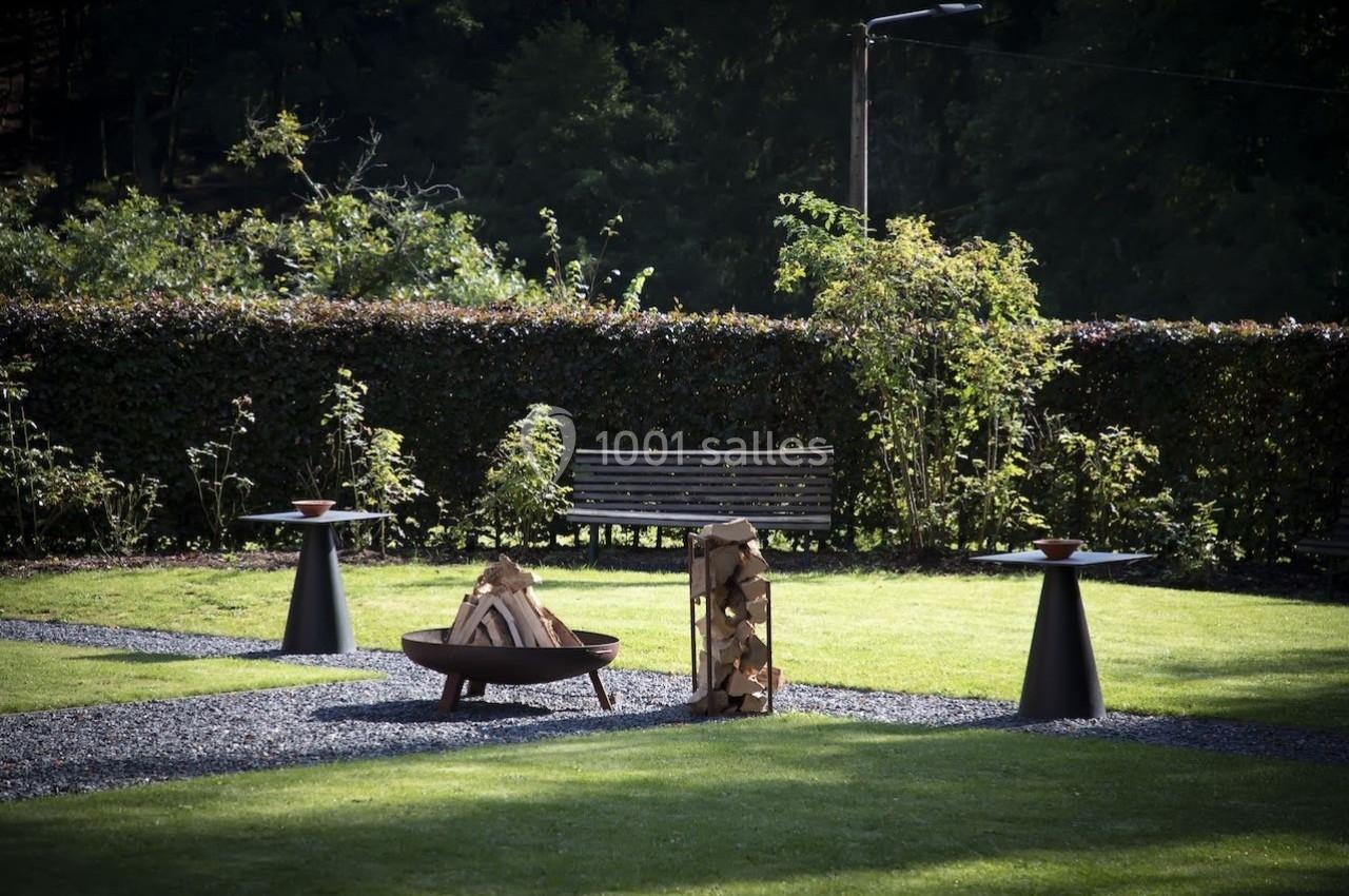 Espace extérieur avec brasero, bûches empilées, banc en bois et tables d'appoint sur une pelouse entourée de haies.