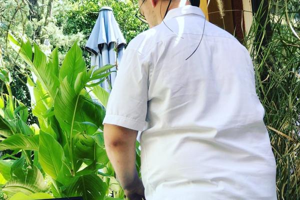 Un homme en chemise blanche vu de dos, debout près de plantes vertes dans un jardin avec un parasol en arrière-plan.