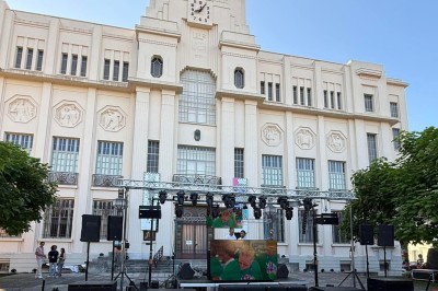Scène extérieure équipée de matériel audio et éclairage, entourée de barrières, sur une place publique.