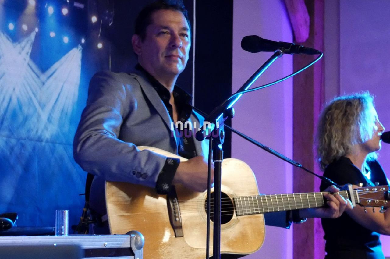 Un musicien joue de la guitare acoustique sur scène, accompagné d'une chanteuse au micro, sous des éclairages colorés.