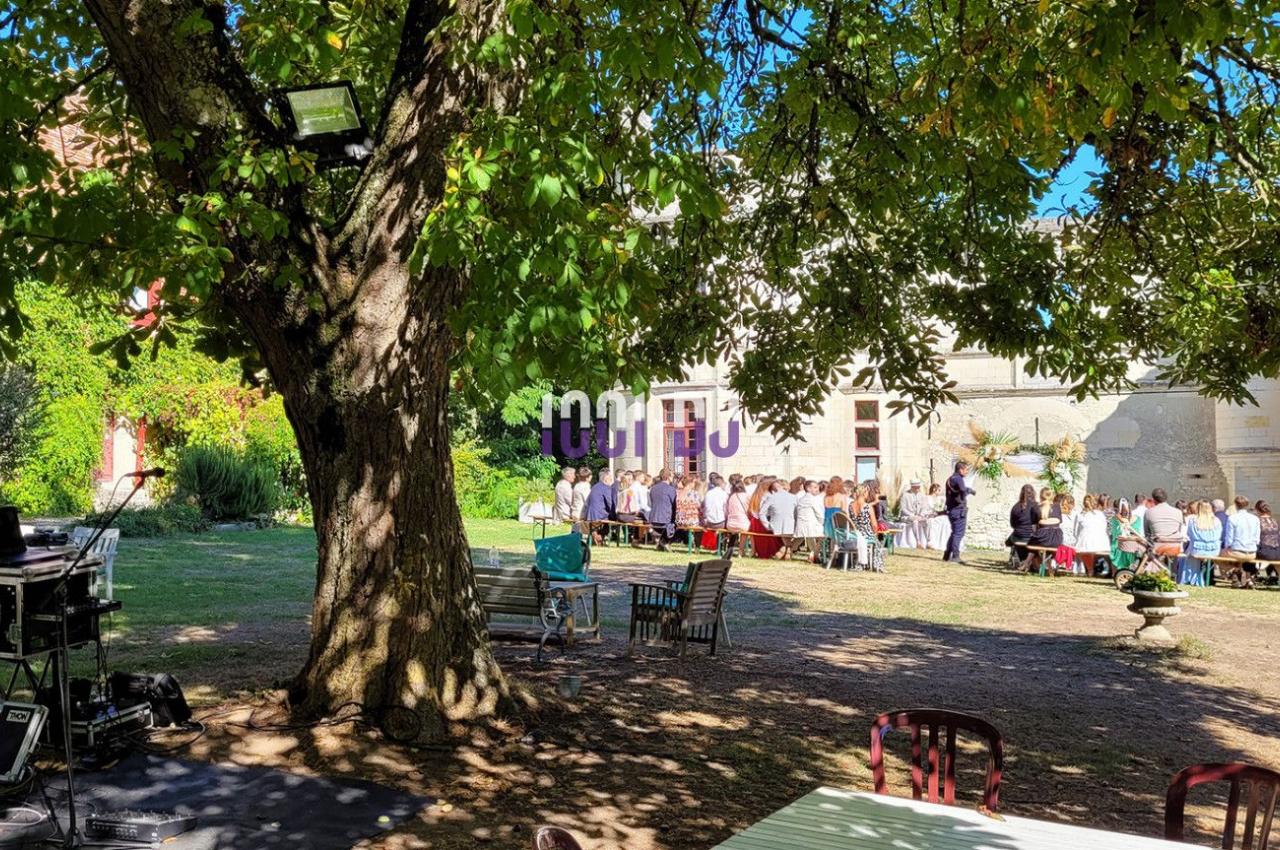 Un groupe de personnes assises à l'extérieur sous des arbres, près d'un bâtiment, avec du matériel audio visible au premier…