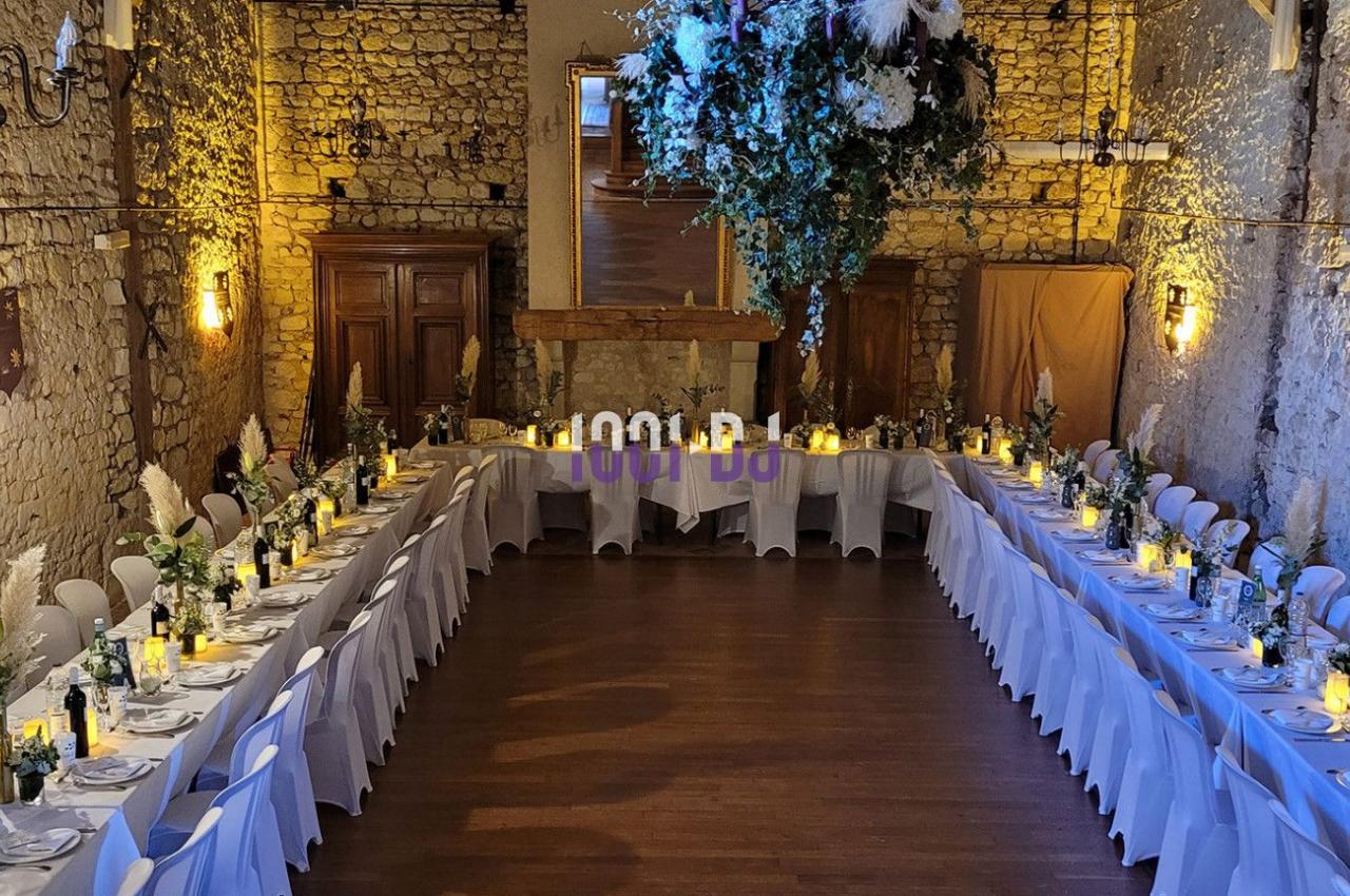 Salle en pierre avec deux longues tables dressées pour un repas, décorée de fleurs suspendues et éclairée par des bougies.