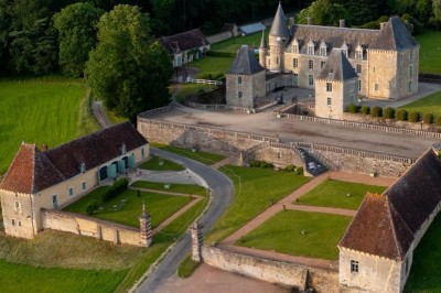 Château éclairé de nuit avec des lumières colorées, entouré de buissons et de tentes blanches sous un ciel nuageux.