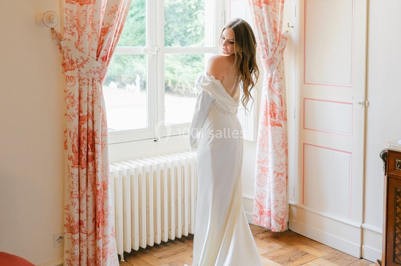 Femme en robe blanche dos nu regardant par-dessus son épaule devant une fenêtre encadrée de rideaux à motifs.