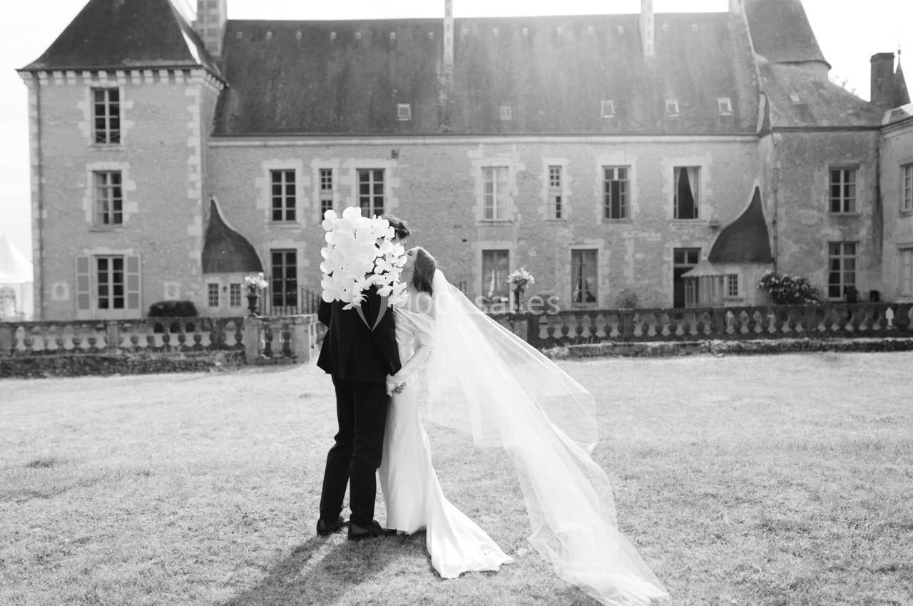 Un couple de mariés s'embrasse dans un jardin devant un bâtiment ancien, avec une longue traîne de voile.