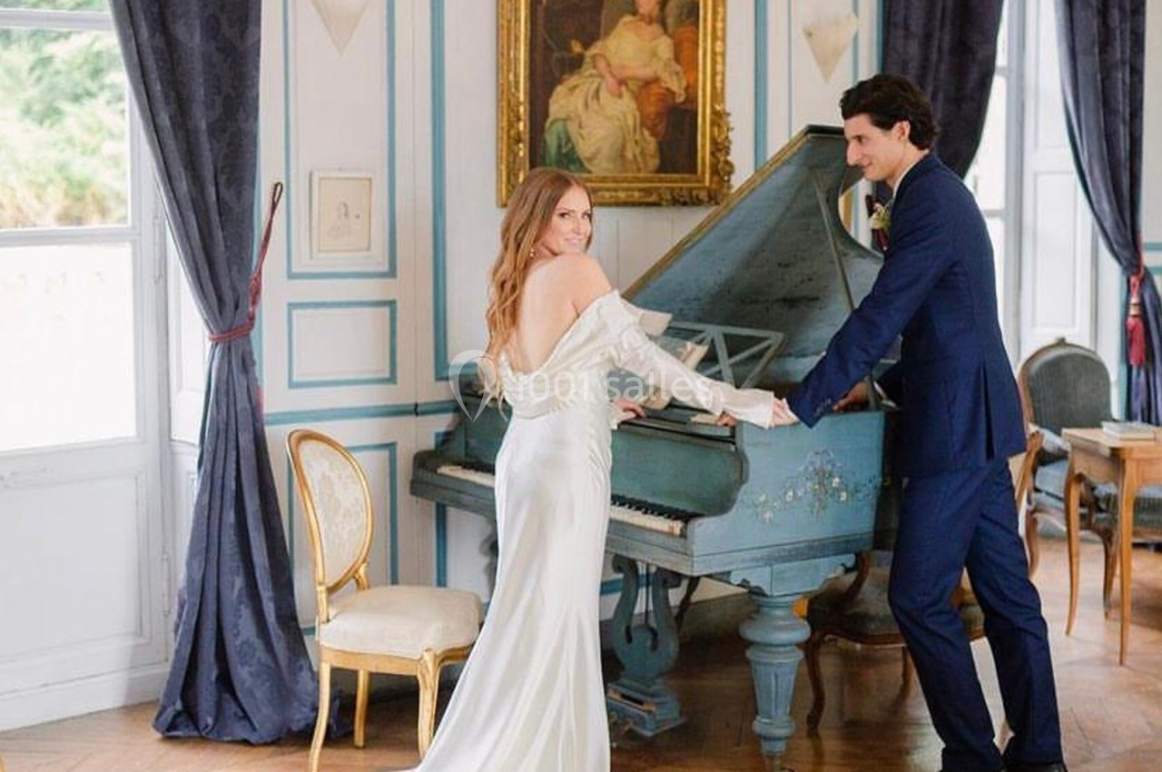 Une femme en robe blanche et un homme en costume posent près d'un piano bleu dans une pièce élégante.