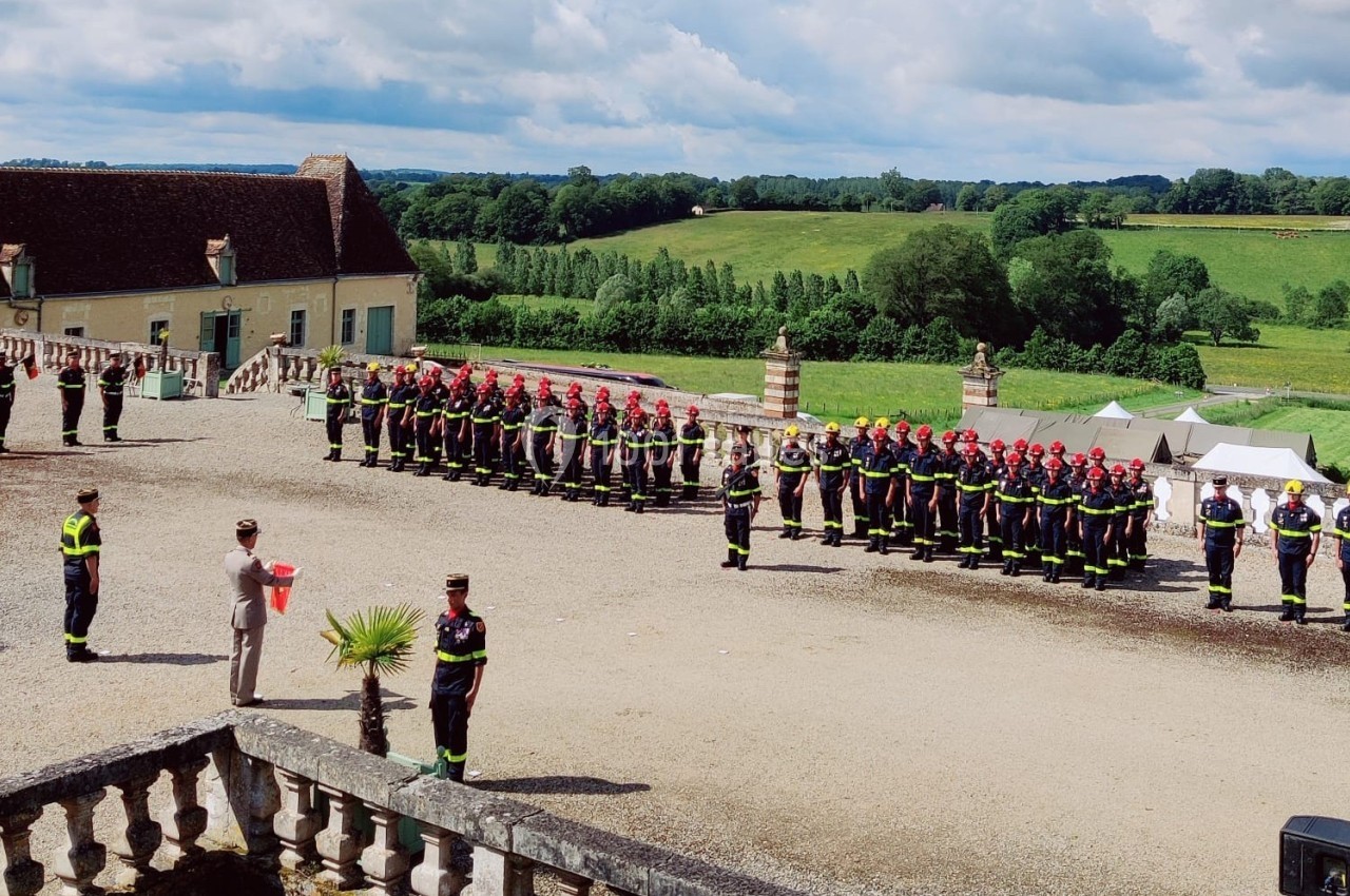Des pompiers en uniforme alignés lors d'une cérémonie en extérieur, avec un bâtiment ancien et un paysage verdoyant en…