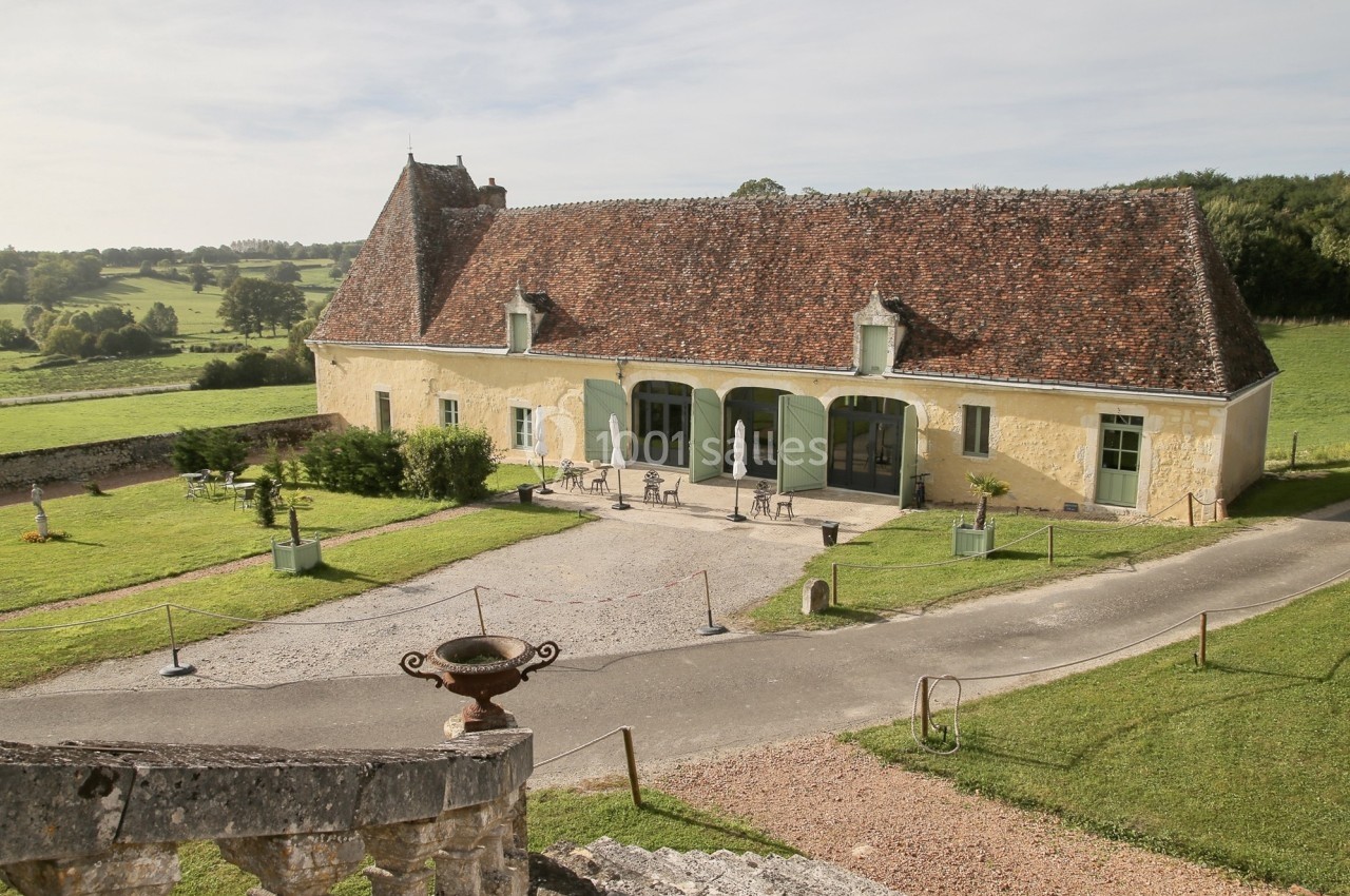 Bâtiment ancien avec toit en tuiles, entouré de verdure et d'une cour gravillonnée, en milieu rural.
