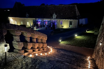 Château éclairé de nuit avec des lumières colorées, entouré de buissons et de tentes blanches sous un ciel nuageux.