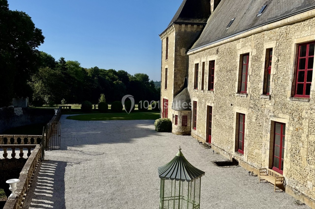 Cour d’un château avec façade en pierre, fenêtres rouges, jardin arboré en arrière-plan et ciel dégagé.