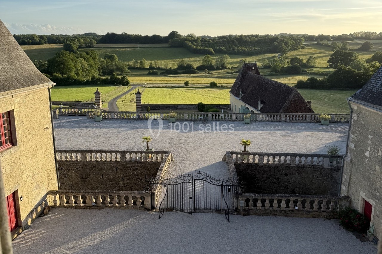 Cour d’un domaine ancien avec portail en fer forgé, bâtiments en pierre et vue sur des champs verdoyants au coucher du…