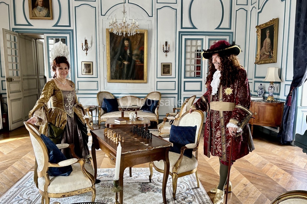 Deux personnes en costumes d'époque dans un salon élégant avec mobilier ancien et échiquier sur une table.