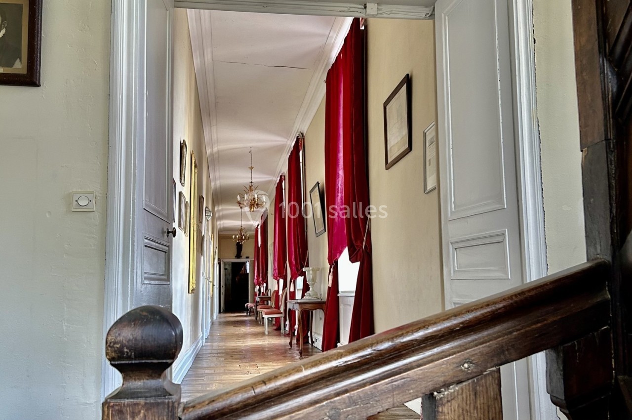 Vue d'un couloir lumineux avec parquet, murs clairs, rideaux rouges et mobilier classique, capté depuis un escalier en bois.