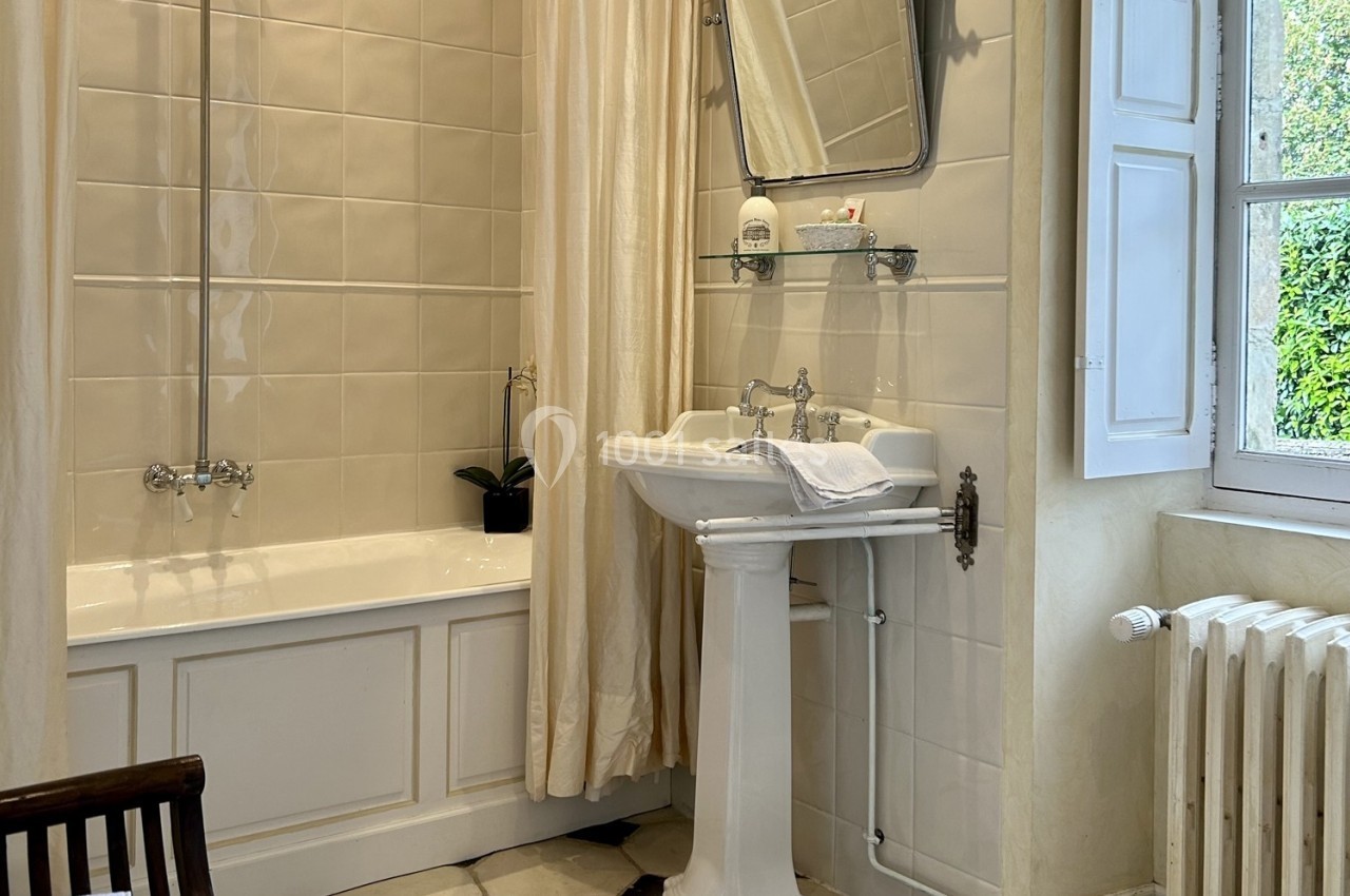 Salle de bain avec baignoire, rideau beige, lavabo sur pied, miroir mural et fenêtre donnant sur un jardin.