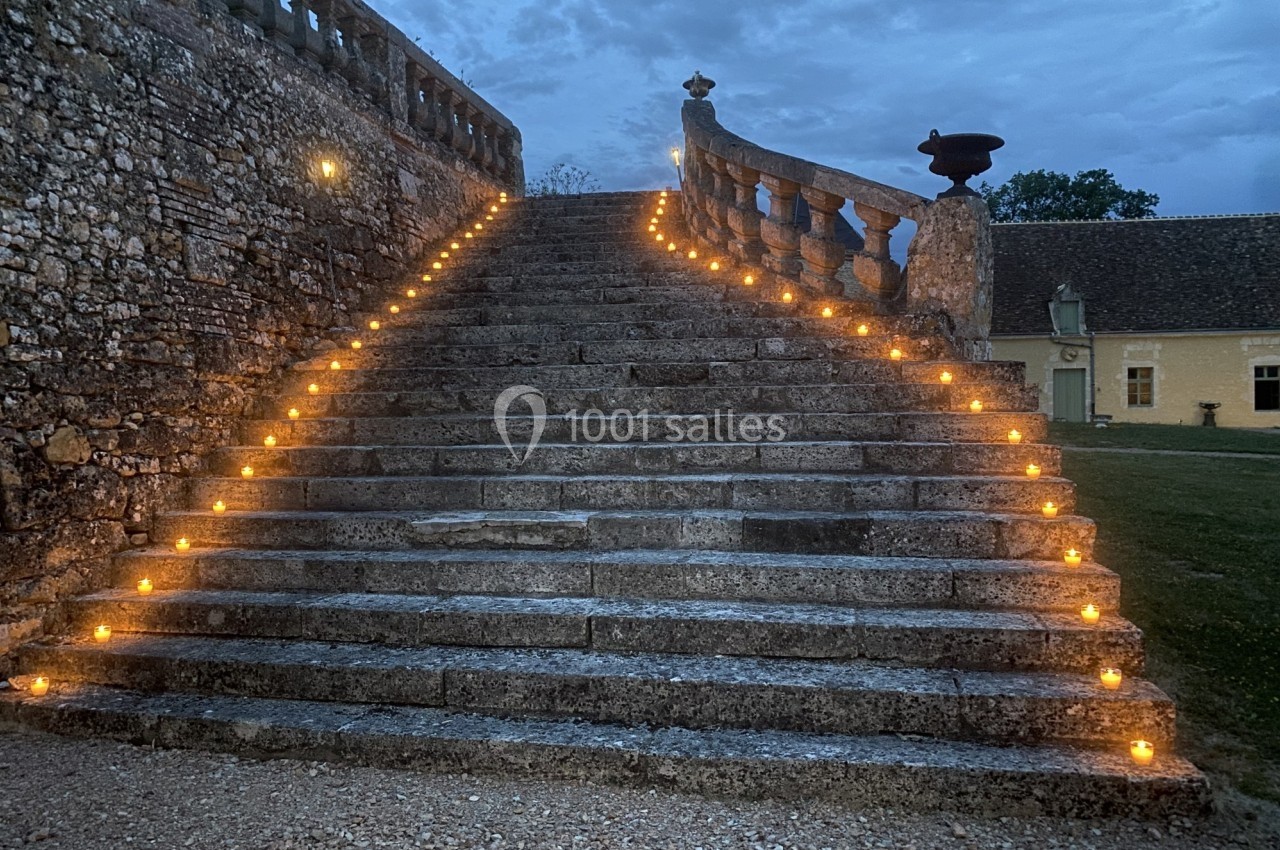 Escalier en pierre éclairé par des bougies alignées, menant à une balustrade, sous un ciel nuageux au crépuscule.