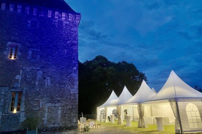 Château éclairé de nuit avec des lumières colorées, entouré de buissons et de tentes blanches sous un ciel nuageux.