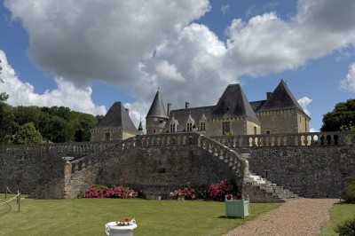 Château éclairé de nuit avec des lumières colorées, entouré de buissons et de tentes blanches sous un ciel nuageux.
