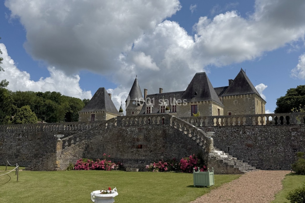 Château en pierre avec tours et toits en ardoise, entouré d'un jardin fleuri et d'un mur en pierre sous un ciel nuageux.