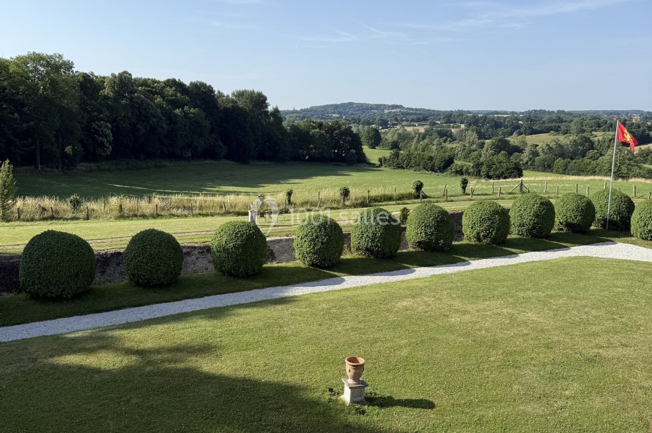 Vue sur un jardin avec pelouse, haies taillées en boules, allées gravillonnées et paysage vallonné en arrière-plan.