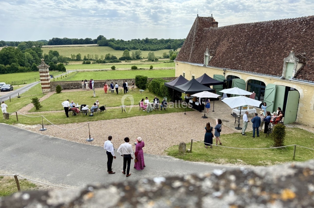 Personnes rassemblées dans un jardin devant un bâtiment ancien, avec des tables, parasols et une vue sur la campagne.