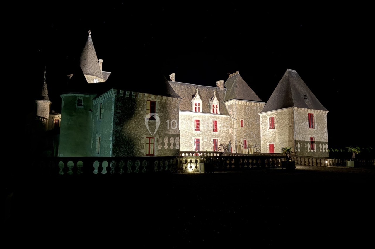 Château en pierre éclairé la nuit, avec des tours et des fenêtres rouges, vu depuis une cour sombre.
